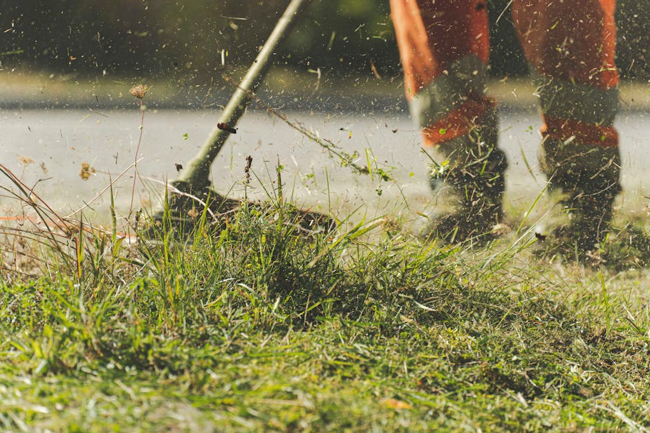 Close-up view of a garden during maintenance work, with a person trimming overgrown grass and weeds using a rake or similar tool. The scene depicts wet soil and scattered plant debris, indicating rece