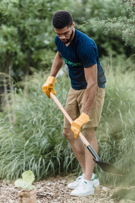 Close-up view of a garden during maintenance work, with a person trimming overgrown grass and weeds using a rake or similar tool. The scene depicts wet soil and scattered plant debris, indicating rece